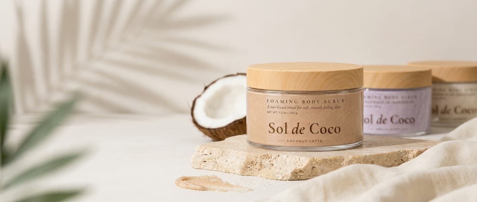 A jar of Sol de Coco Coconut Latte on a travertine slab beside half a coconut, with two more jars and cream linen behind and a palm shadow across the wall.