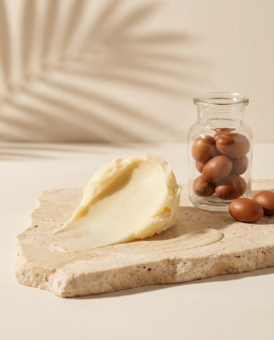 A thick swirl of ivory shea butter on travertine, beside a glass jar of shea nuts, lit softly with a palm shadow behind.