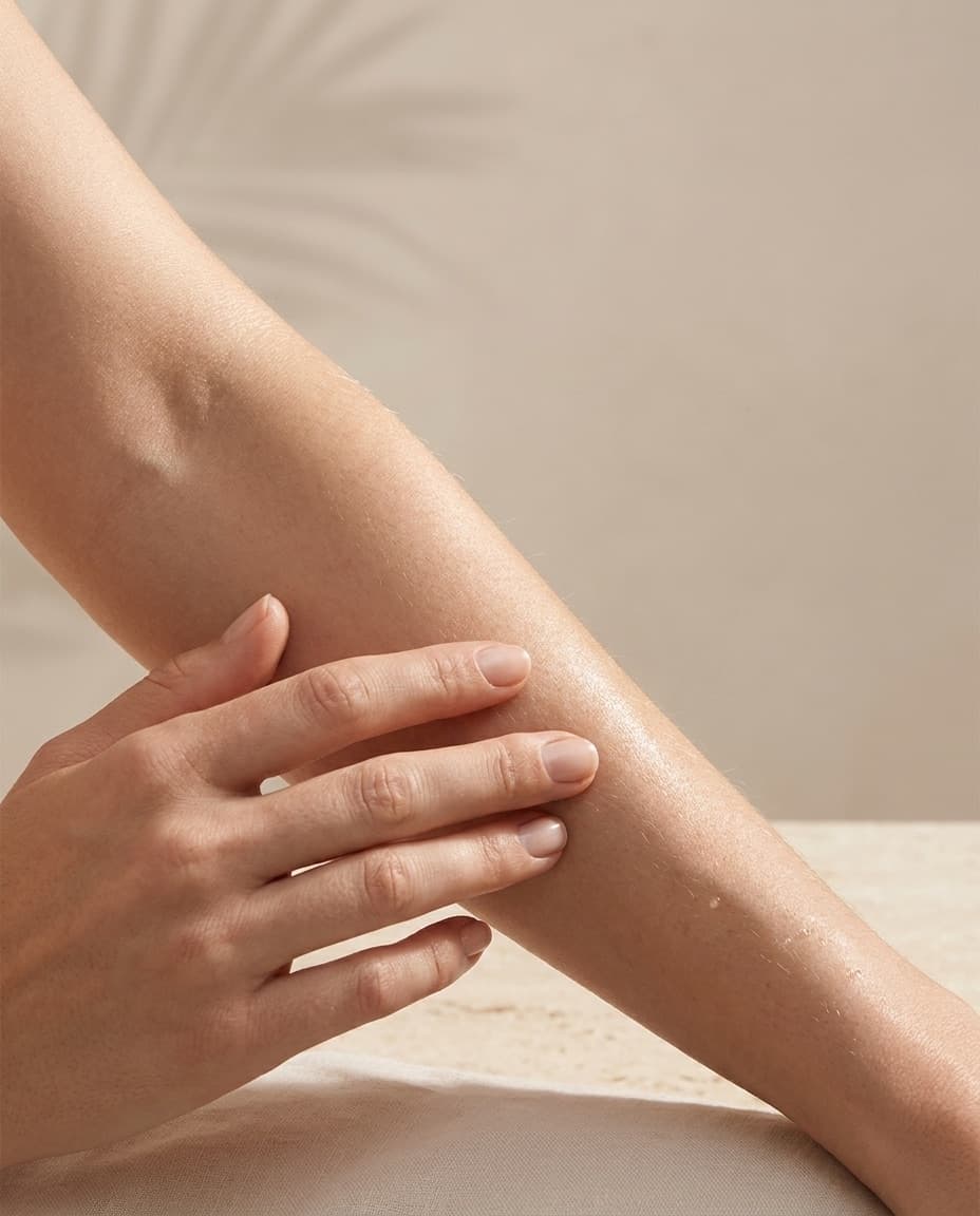 A hand smoothing oil along a forearm after rinsing, resting on travertine with a palm shadow on the wall behind.